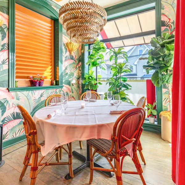 A vibrant restaurant interior with tropical wallpaper, rattan chairs, and a white-clothed table set for dining.