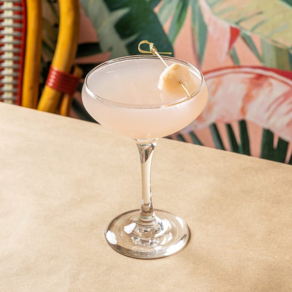 A lychee martini cocktail garnished with a single lychee on a skewer, against a tropical background.
