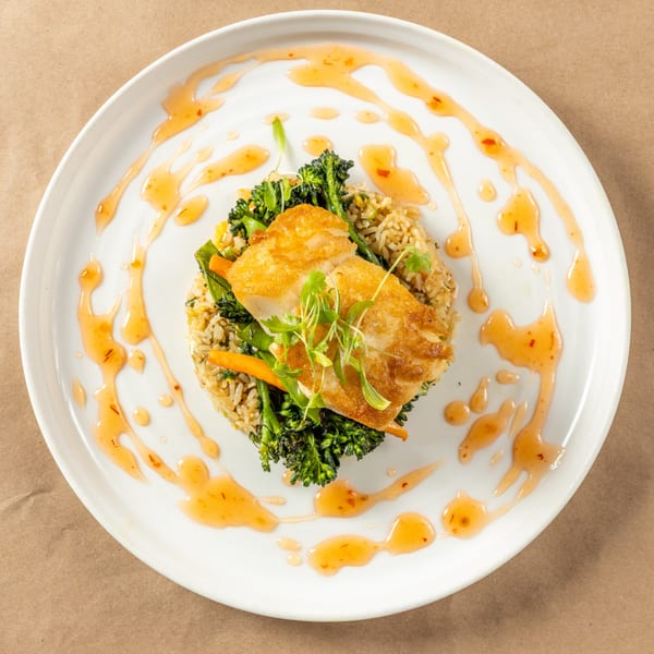 Pan-seared fish over rice and broccolini, garnished with microgreens and sweet chili sauce.