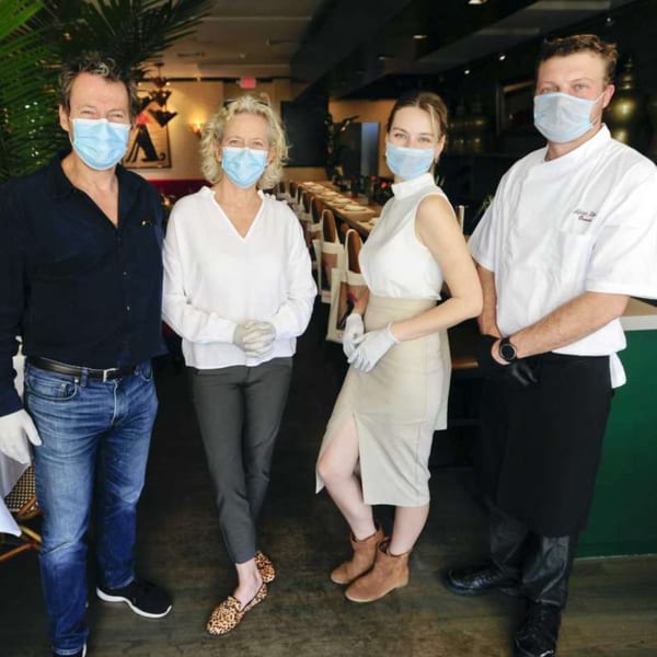 Four restaurant staff members, including a chef, wearing face masks and gloves inside a dining room.
