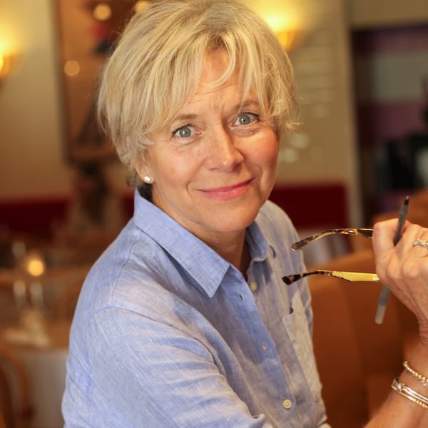 A smiling woman with short blonde hair, wearing a blue shirt, holding glasses and a pen.