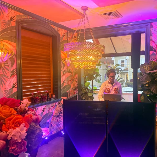 A DJ with headphones and a laptop behind a booth, bathed in pink and purple light.