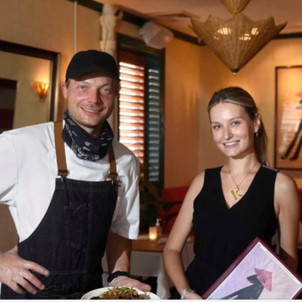 Smiling chef in apron and woman holding a menu stand in a restaurant dining room.