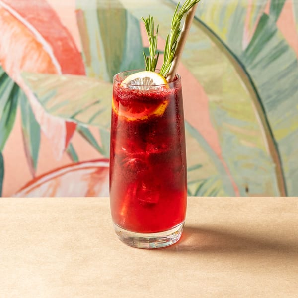 Orienta Restaurant's red cocktail with lemon, rosemary, and a straw against a tropical background.