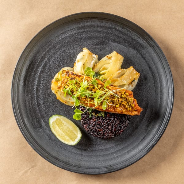Asian-fusion dish with salmon, black rice, roasted fennel, microgreens, and a lime wedge on a black plate.