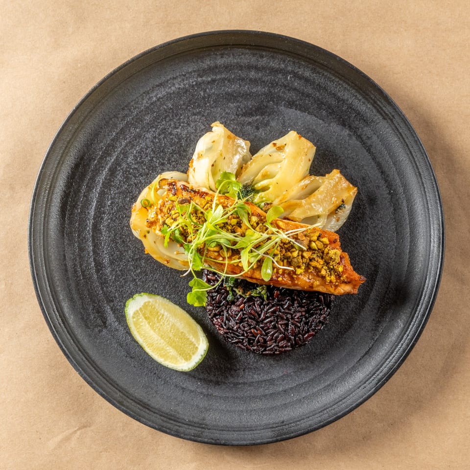 Asian-fusion dish with salmon, black rice, roasted fennel, microgreens, and a lime wedge on a black plate.